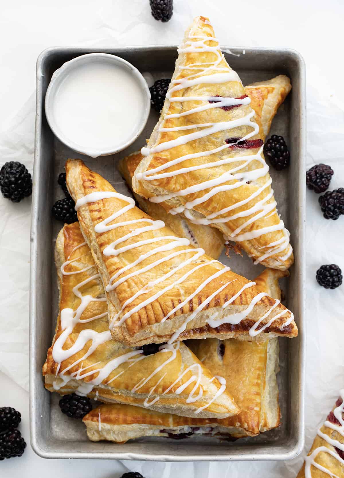 Blackberry Cream Cheese Turnovers on a Pan with Glaze and a Few Blackberries on a White Counter.