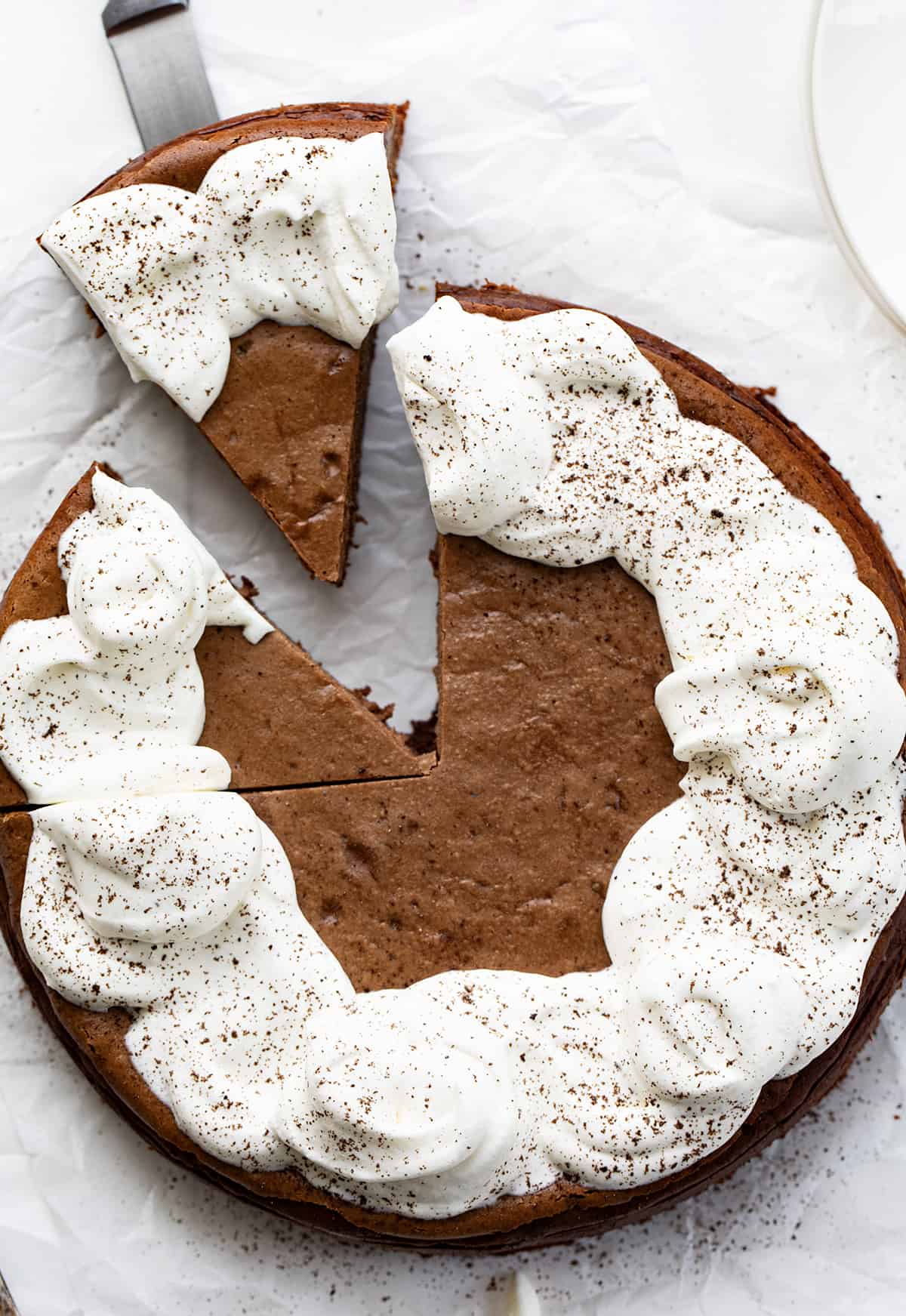 Whole Brownie Bottom Chocolate Cheesecake on White Parchment with Homemade Whipped Topping and Dusted with Chocolate with One Piece Being Removed.