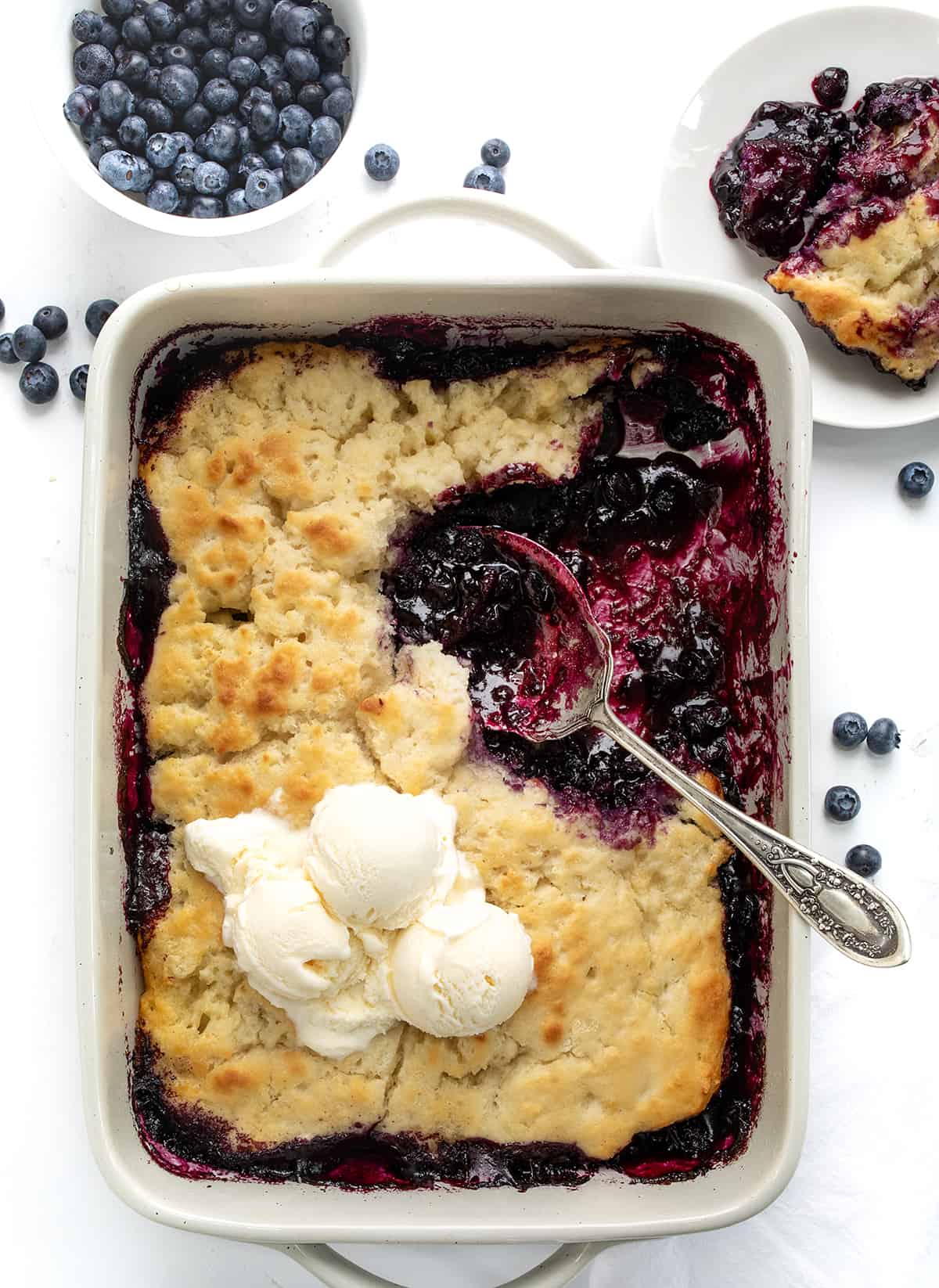Pan of Butter Swim Biscuit Blueberry Cobbler with Ice Cream and a Plated Piece Next to Fresh Blueberries from Overhead.