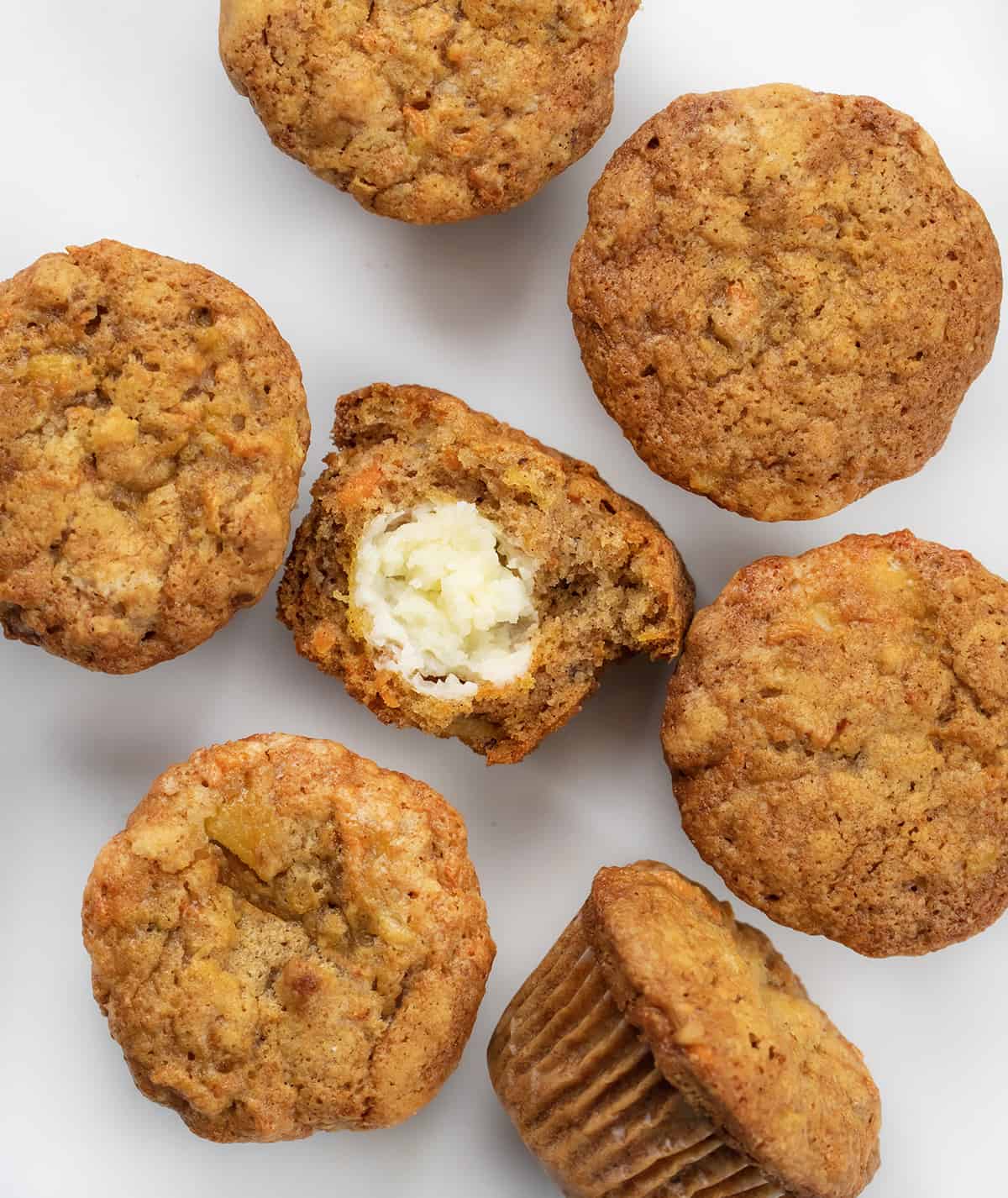 Cream Cheese Filled Carrot Cake Cupcakes on a White Counter With One Halved Showing Cream Cheese Inside.