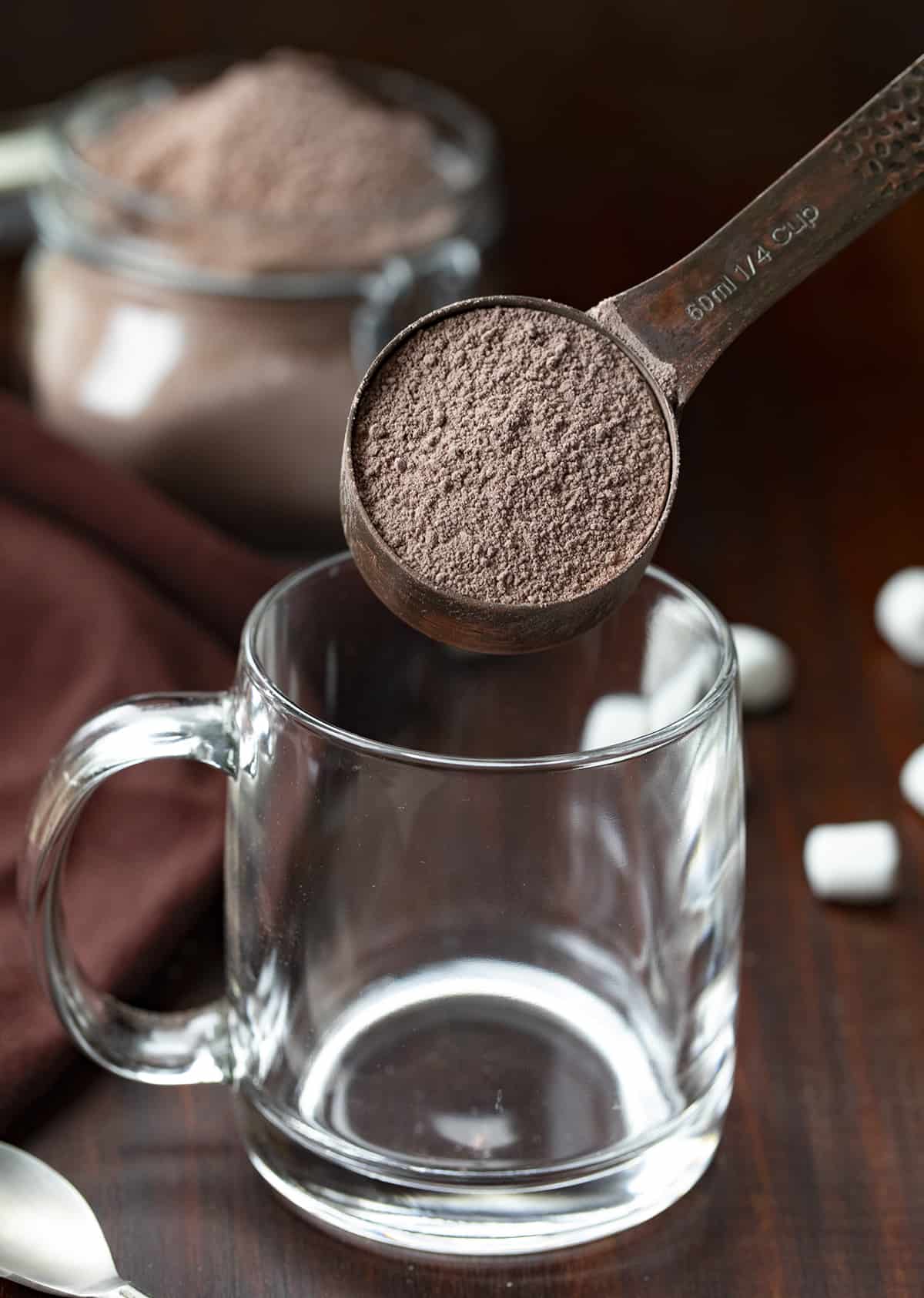 A 1/4 cup measuring scoop adding hot cocoa mix to an empty glass.