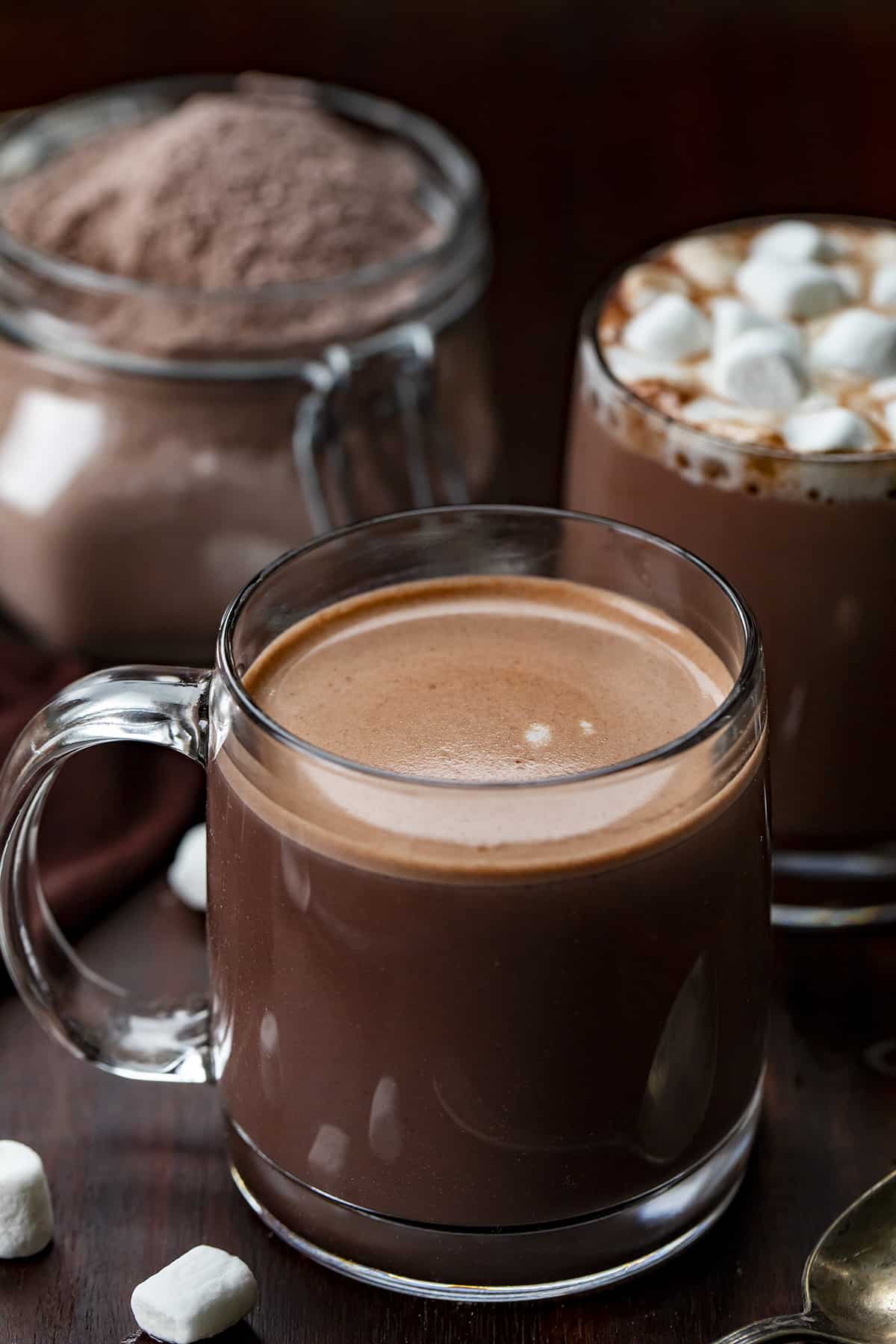 Glasses of prepared hot chocolate made from a homemade hot cocoa mix with the jar of the mixture in the background. 