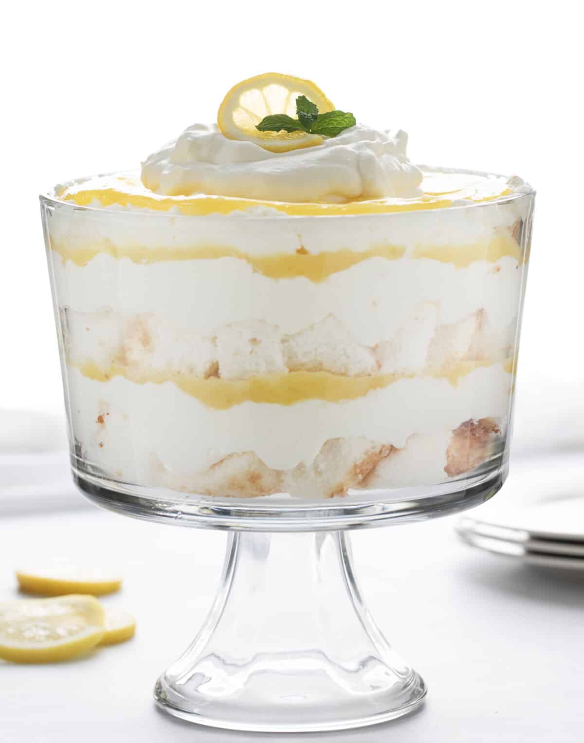 A Lemon Cream Trifle in a Glass Trifle Dish on a White Counter with Lemons and Plates.